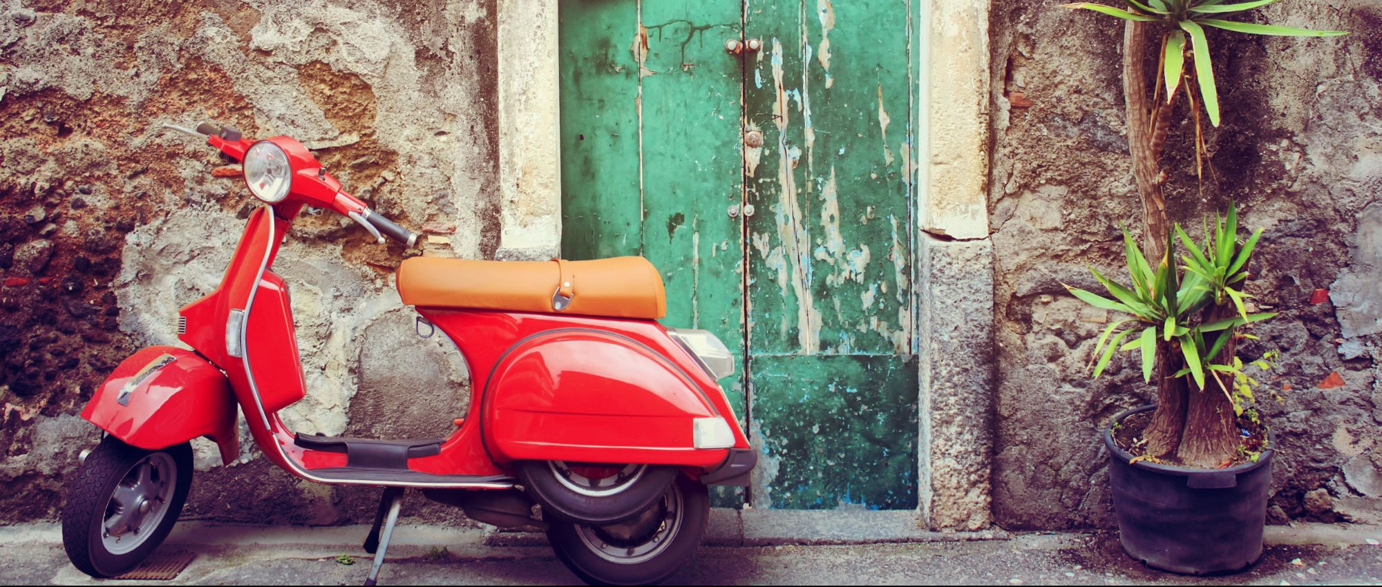 Klassische rote Vespa vor einer alten Steinwand mit grüner Holztür – Sinnbild italienischer Lebensfreude und Nostalgie.